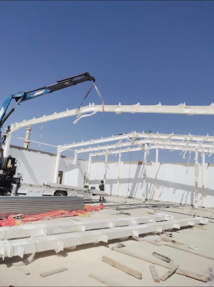 Crane lifting a white curved steel roof beam into position on an E&M site